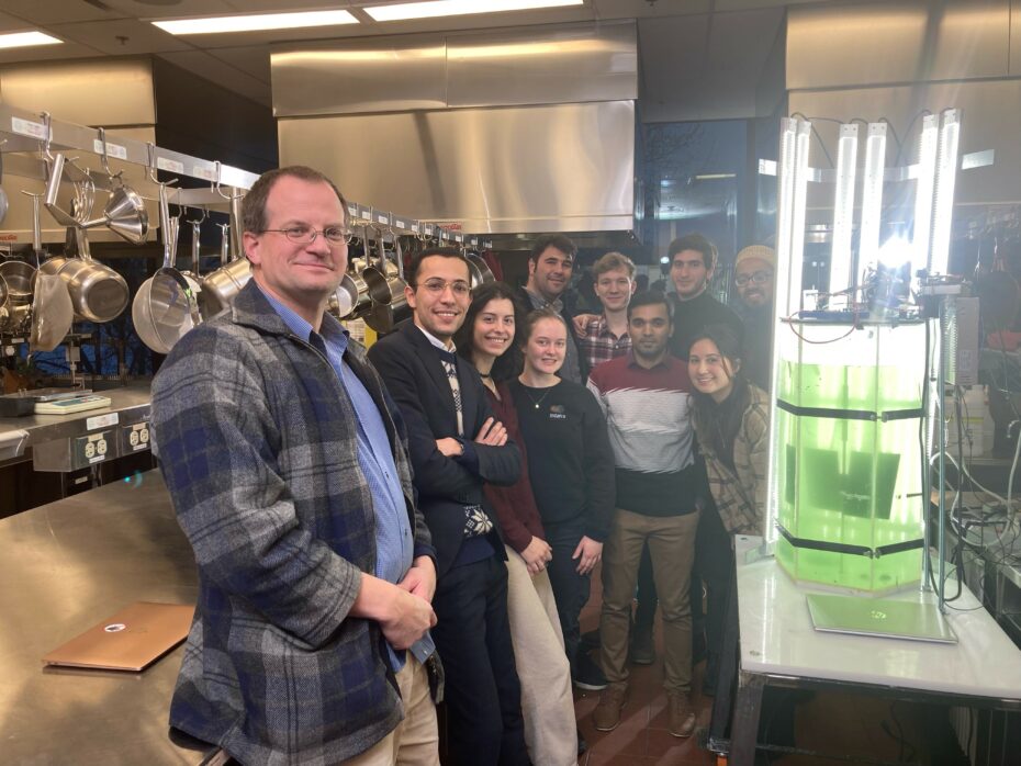 Professor and students in laboratory with a brightly lit tank of pale green liquid