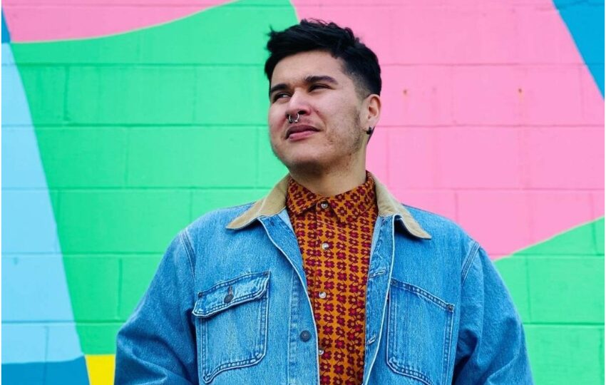 Billy-Ray Belcourt wearing a jean jacket and standing in front of a mural wall of pastel colours