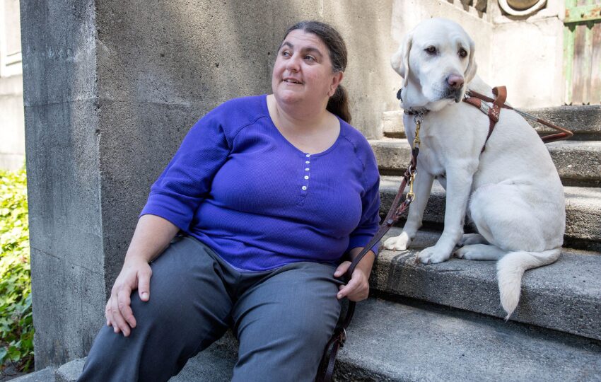 Lucy Greco, wearing purple shirt and grey pants, poses on concrete steps with her white guide dog, Frances