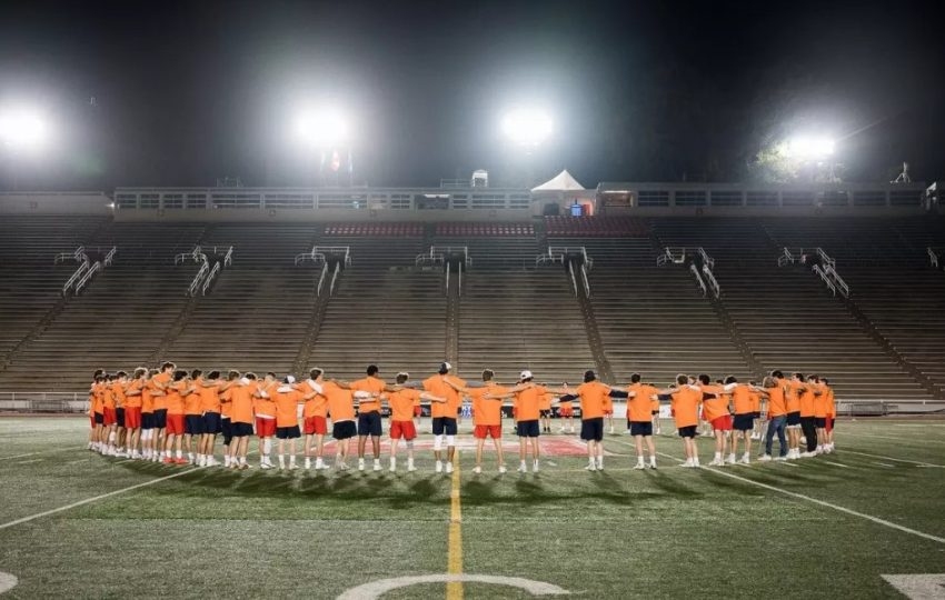 Lacrosse players on McGill's field standing in a circle and wearing orange shirts